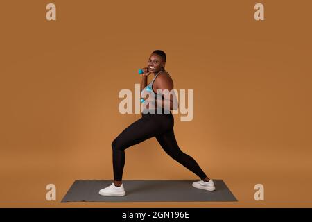 Strength Training Concept. Happy curvy black lady doing lunges exercise with dumbbells on brown studio background, standing on yoga mat, looking at ca Stock Photo