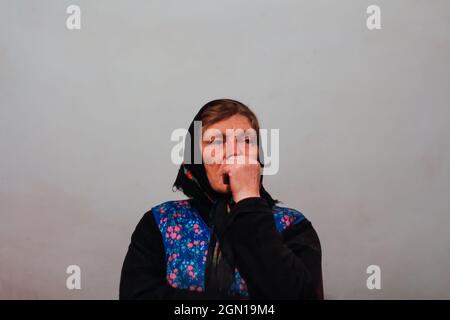 Defocus portrait of 60s russian grandmother senior old woman seating indoors. Old women in shawl. Sad thinking person. White dirty background. Out of Stock Photo