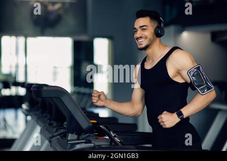 Handsome Muscular Arab Man In Wireless Headphones Jogging On Treadmill At Gym Stock Photo