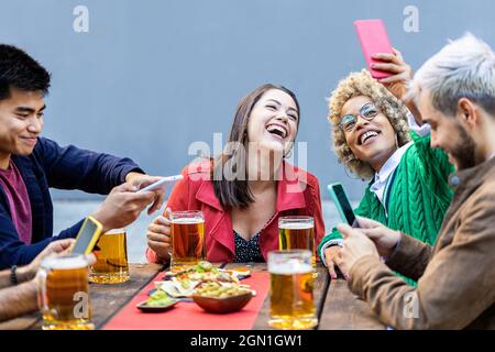 Group of happy millennial people using smartphone while drinking beer at terrace Stock Photo