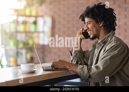 Black Freelancer Guy Talking On Cellphone While Working With Laptop At Cafe Stock Photo