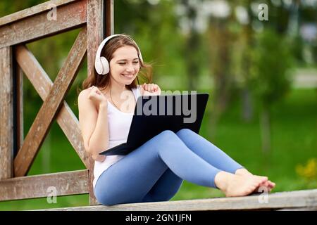 Happy woman in cordless headphones has chat in laptop outdoors. Stock Photo