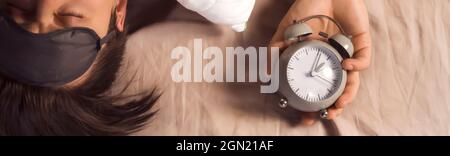Hands of a young man from under the blankets hold an alarm clock in gray.  Stock Photo