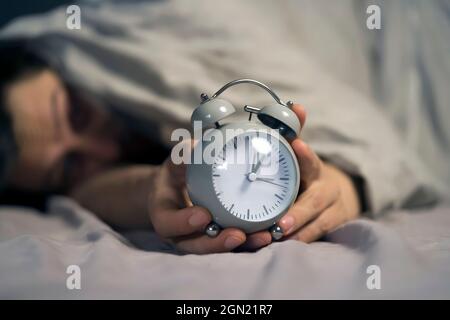 Hands of a young man from under the blankets hold a retro vintage alarm clock. Stock Photo