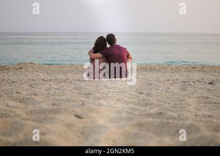 Young couple man and woman are sitting at the beach and looking at the sea. Back Stock Photo