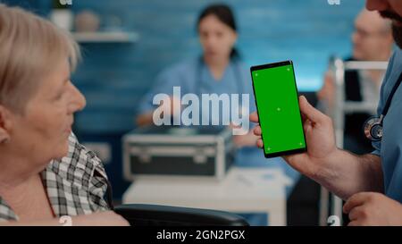 Close up of nurse vertically showing phone with green screen to senior patient living in nursing home. Medical specialist holding smartphone with chroma key and isolated mockup template Stock Photo