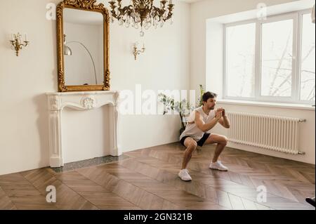 Strong Athletic Fit Man in T-shirt and Shorts is Doing Squat Exercises at Home in His Spacious and Bright Apartment Stock Photo