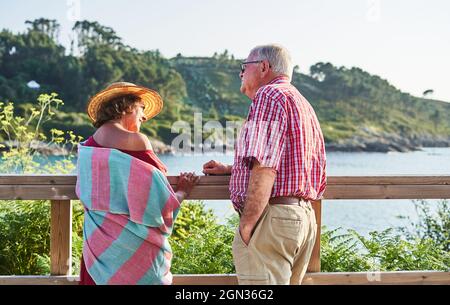 Back view of senior couple standing close to each other looking at camera and enjoying view of sea in sunny day Stock Photo