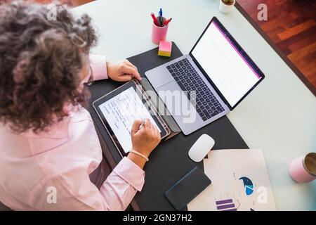 From above of crop anonymous female entrepreneur with tablet and netbook working at desk with graphics on paper sheets Stock Photo