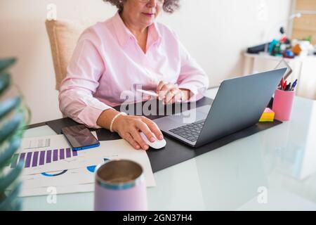 Crop senior female entrepreneur with tablet and netbook working at desk with graphics on paper sheets Stock Photo
