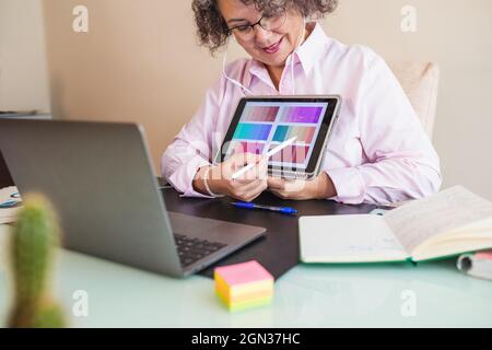 Crop senior female in earphones touching screen on tablet while pointing at color palette and speaking during video chat on netbook in office Stock Photo