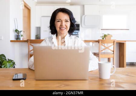 Happy mature female freelancer browsing on Internet on netbook working on new project while sitting at table at home Stock Photo