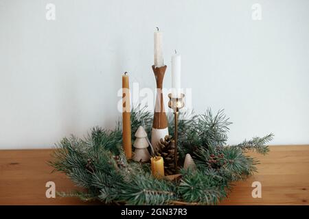 Stylish candles in christmas wreath  with pine cones and tree decor on wooden table on background of white wall in festive room. Christmas advent. Atm Stock Photo