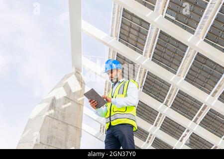 Low angle of middle aged Hispanic male engineer in uniform with tablet standing looking at screen under solar power station Stock Photo