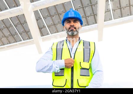 Mature ethnic male contractor in hardhat and vest standing looking away near solar power station Stock Photo