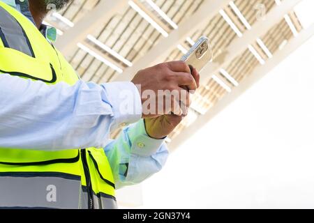 Cropped unrecognizable foreman in waistcoat browsing on smartphone while standing near solar power station Stock Photo