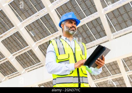 Low angle of middle aged Hispanic male engineer in uniform with tablet standing looking away under solar power station Stock Photo