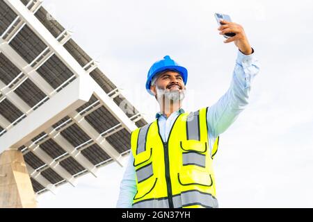 Low angle of cheerful mature ethnic male supervisor in helmet and waistcoat taking self portrait on cellphone against modern construction in sunlight Stock Photo