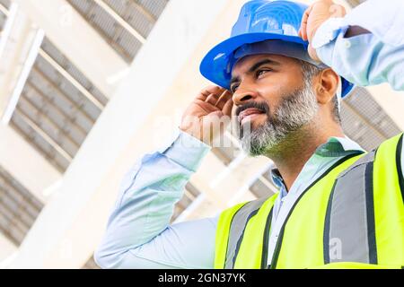 From below of mature ethnic male supervisor in vest putting on hardhat while looking away standing near solar power station Stock Photo