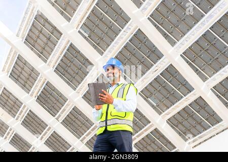 Low angle of happy middle aged Hispanic male engineer in uniform with tablet standing looking away under solar power station Stock Photo