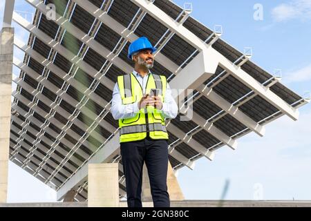 Smiling middle aged Hispanic foreman in in hardhat and waistcoat browsing on smartphone looking away while standing near solar power station Stock Photo