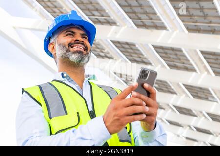 From below happy middle aged Hispanic foreman in in hardhat and waistcoat browsing on smartphone looking at screen while standing near solar power sta Stock Photo