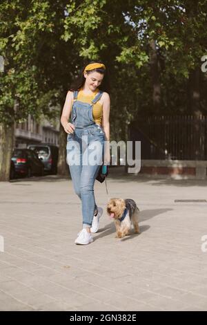 Full body of positive female owner in casual denim overall walking with Yorkshire Terrier on leash Stock Photo
