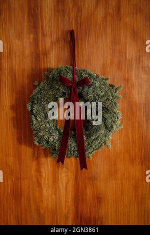 Decorative coniferous wreath with red bow hanging on wooden wall in room during Christmas Stock Photo