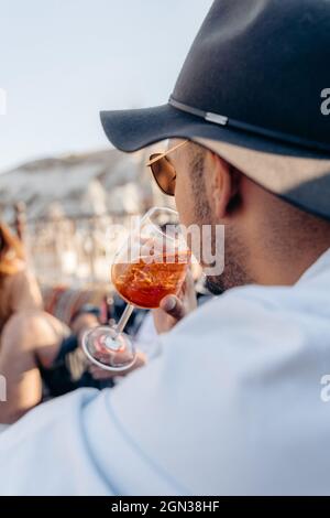 Side view of young bearded male in sunglasses and stylish hat sipping cold refreshing cocktail while relaxing on terrace Stock Photo