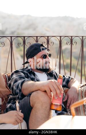 Adult bearded male in stylish sunglasses sitting on couch with bottle of beer near crop friend Stock Photo