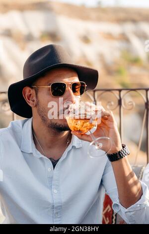 Young bearded male in sunglasses and stylish hat sipping cold refreshing cocktail while relaxing on terrace in Cappadocia, Turkey Stock Photo
