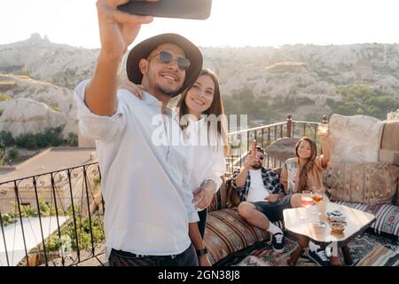 Group of young diverse friends taking selfie on cellphone while drinking cocktails on terrace bar in Cappadocia, Turkey Stock Photo