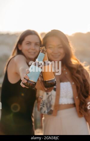 Positive young multiethnic girlfriends smiling brightly and clinking bottles of beer while enjoying pleasant time together at sunset on terrace bar in Stock Photo