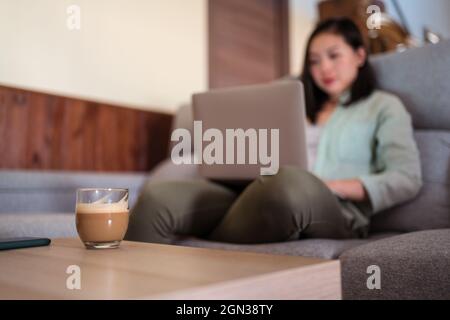 Interested young ethnic female remote employee working on netbook on sofa against coffee and smartphone at home Stock Photo