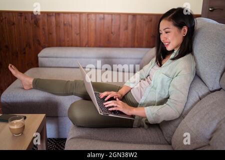 Side view of smiling young ethnic female remote employee working on netbook on sofa against coffee and smartphone at home Stock Photo