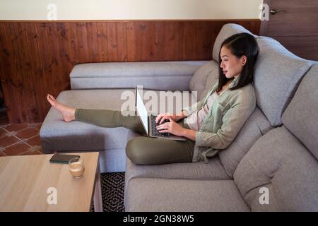 Side view of interested young ethnic female remote employee working on netbook on sofa against coffee and smartphone at home Stock Photo