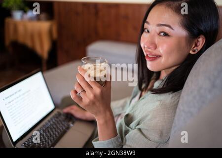 Side view of interested young ethnic female remote employee working on netbook on sofa drinking coffee at home looking at camera Stock Photo