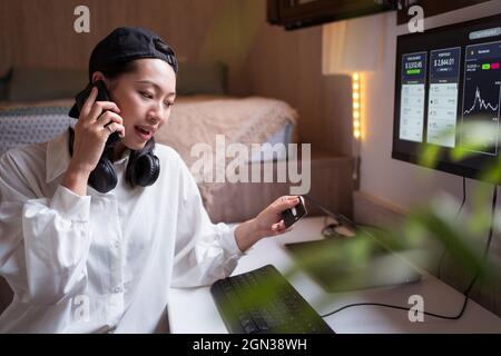 Surprised Asian female trader speaking on smartphone and looking at credit card while working with cryptocurrency statistic Stock Photo