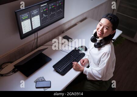 High angle cheerful Asian female working on computer with charts showing dynamic of changes in value of cryptocurrency at convenient workplace Stock Photo