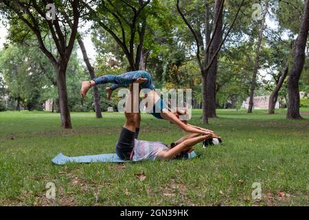 Side view of flexible couple in sportswear practicing acroyoga together on yoga mat on grass against trees in park in daytime Stock Photo