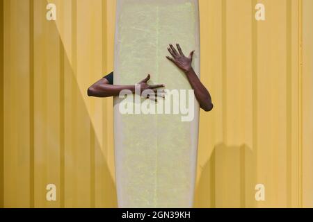 Anonymous ethnic sportsWoman hiding behind longboard surf against yellow ribbed wall with shadow on sunny day Stock Photo