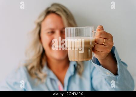 Content adult blond female holding cup of delicious coffee with milk foam on top on light background Stock Photo