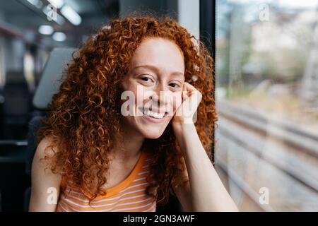 Cheerful young Woman with curly red hair looking at camera during trip on train Stock Photo