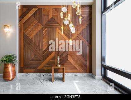 Hall with wood cladding wall decorated with geometrical patterns, tall rounded wooden planter with green bushes, contemporary glass tall chandelier, big window, and white marble floor in modern house Stock Photo