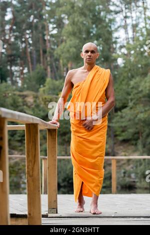 full length view of buddhist monk standing near wooden fence in forest Stock Photo