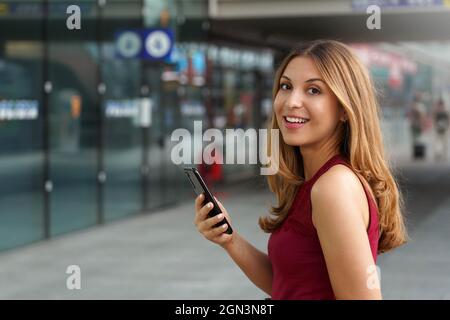 Side view of smiling cute and active Brazilian female traveler passenger standing with smartphone in metro station and looking at camera. Idea for tra Stock Photo