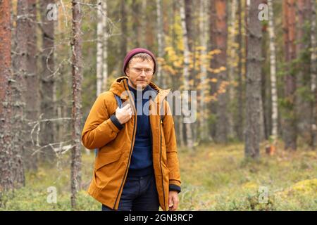 Caucasian man wearing backpack standing over forest background. A man with backpack walking in the autumn forest - Image Stock Photo