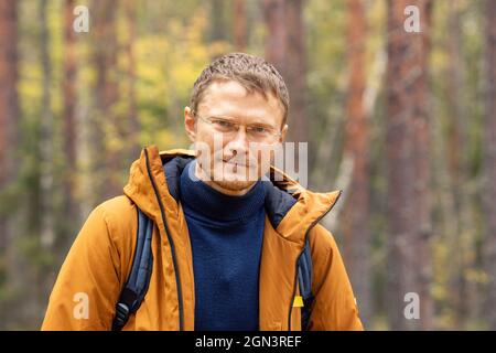 Caucasian man wearing backpack standing over forest background. A man with backpack walking in the autumn forest - Image Stock Photo