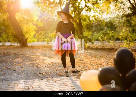 Happy little girl celebrates Halloween outdoors. Child in witch costume and hat with orange and black balloons in autumn park.Children's tricks or Stock Photo