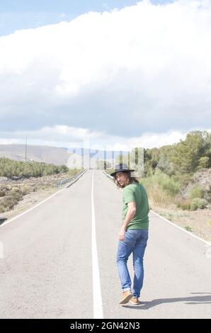 young man in hat walking down a deserted road turns to look Stock Photo